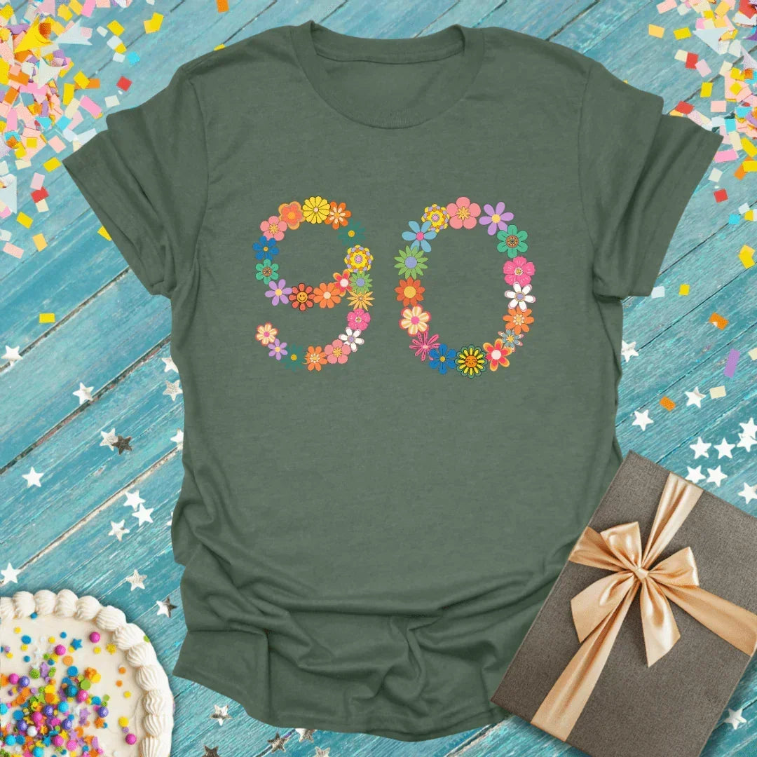 Green 90th birthday tee with floral number design, gift box, and cake on confetti background