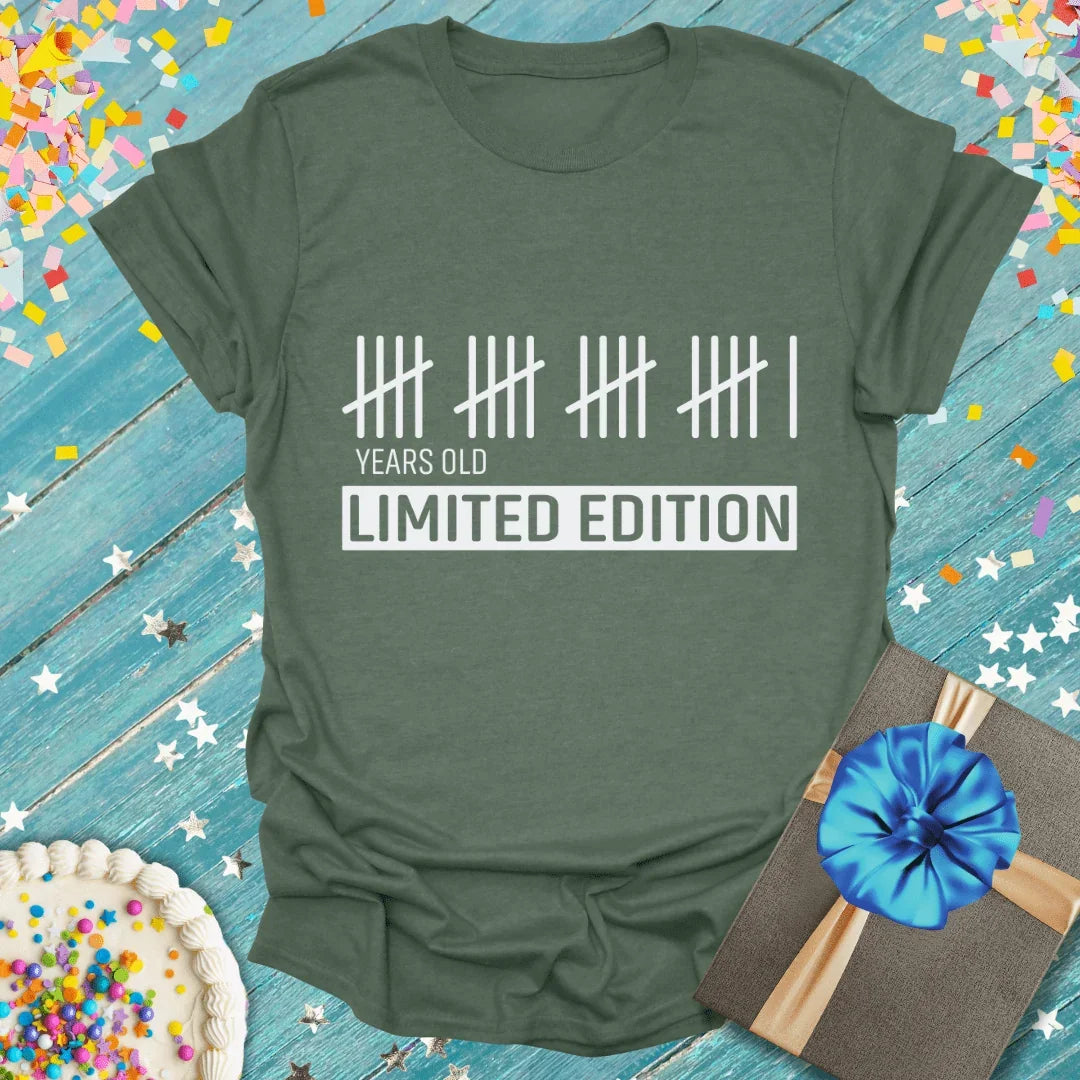 Green birthday tee with tally marks, 'Years Old Limited Edition' text, cake, gift, confetti on blue wood
