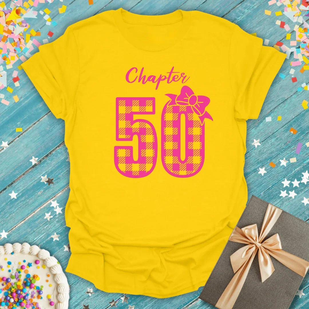 Yellow 'Chapter 50' birthday tee with pink plaid number, gift box, and cake on blue wood