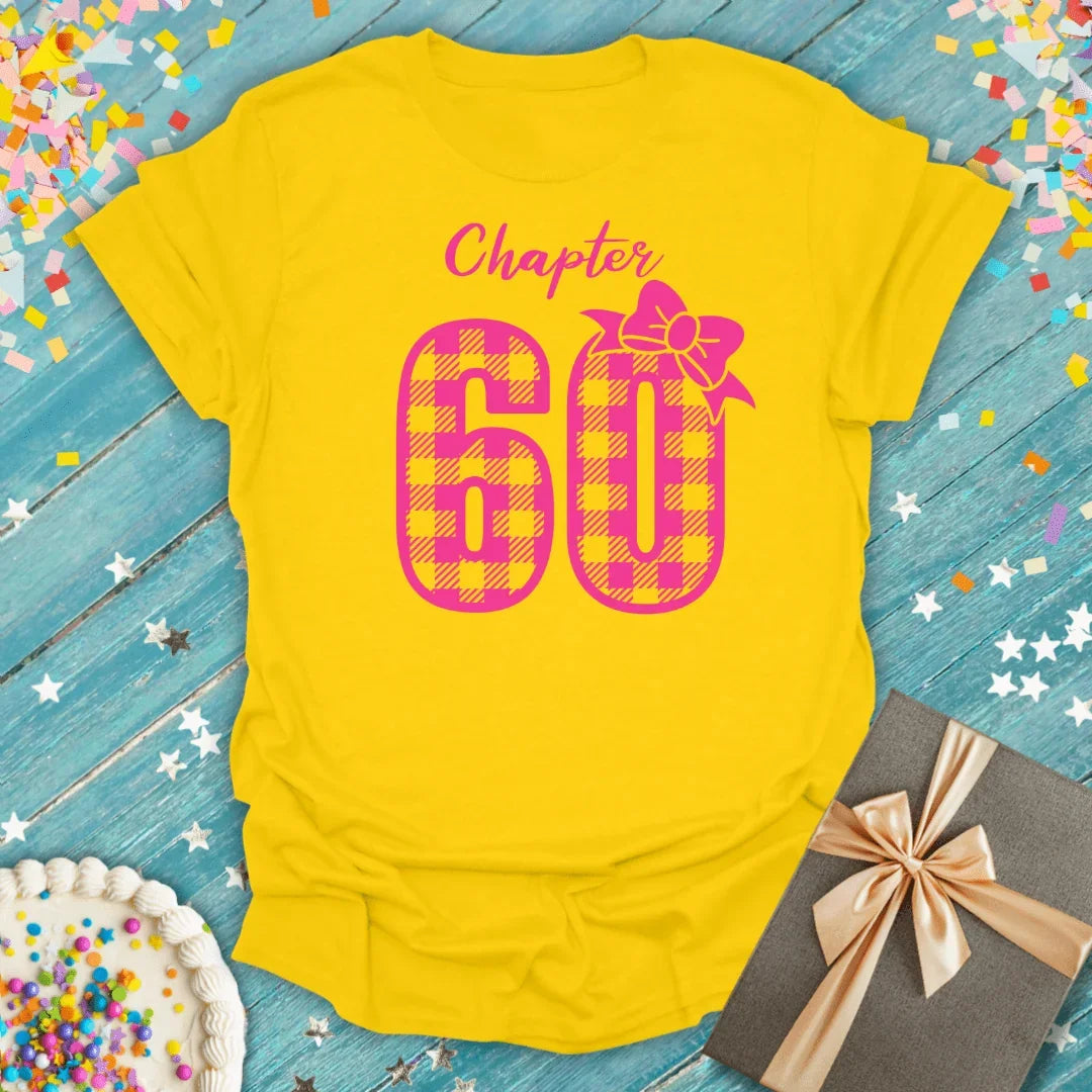 Yellow Chapter 60 birthday tee with pink plaid number, confetti, cake, and gift on blue wood