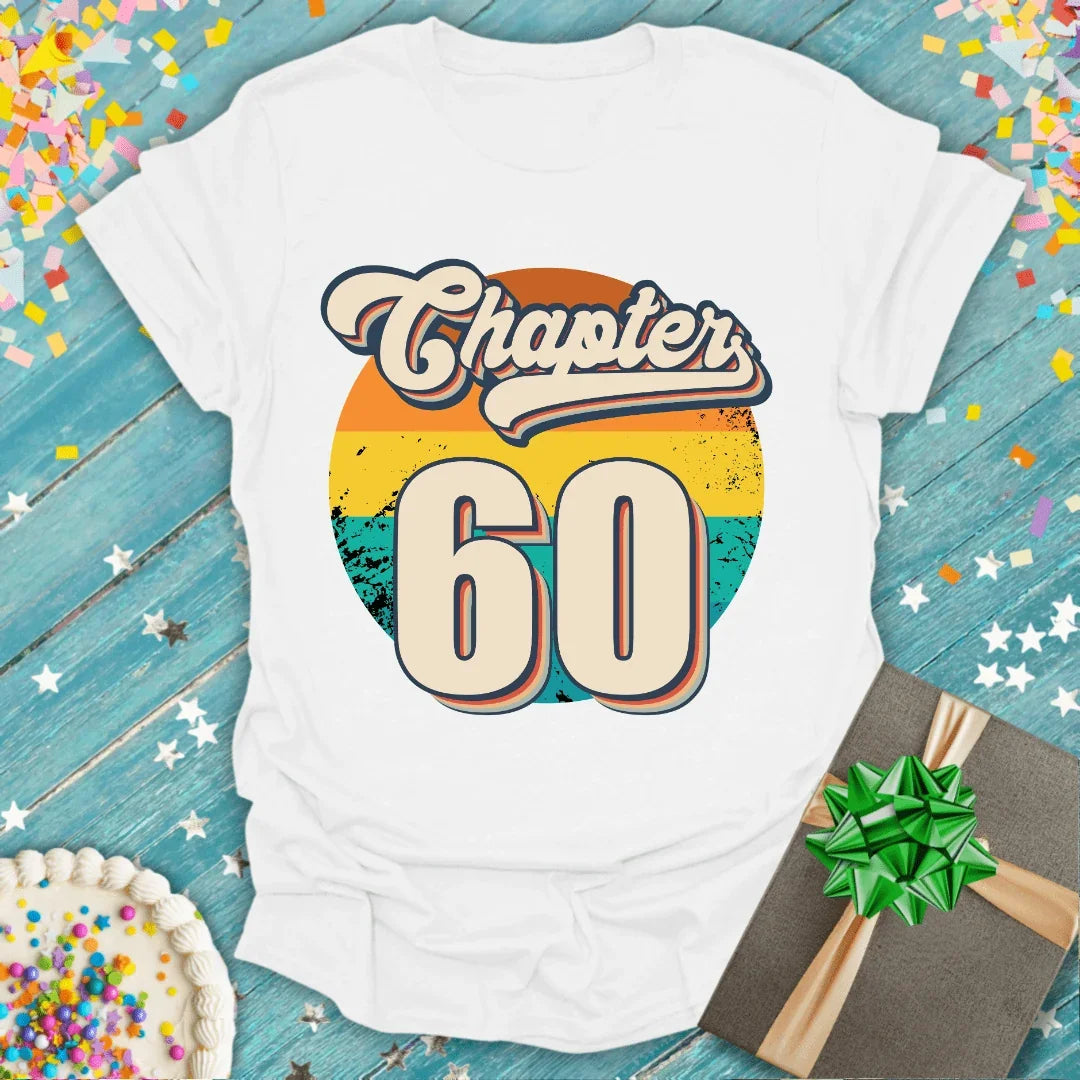White 'Chapter 60' birthday tee with retro sunset design, gift box, and cake on confetti backdrop