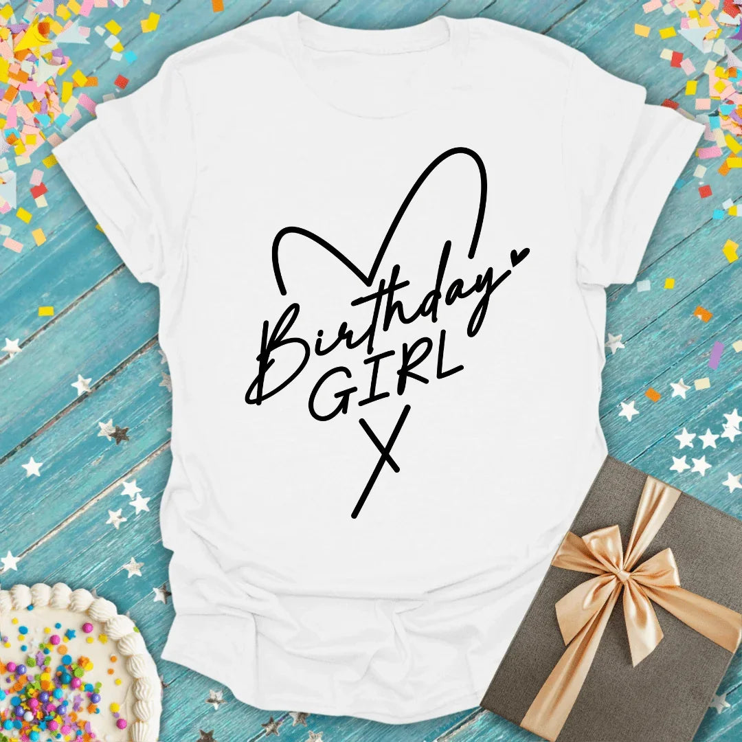 White 'Birthday Girl' t-shirt with heart design, confetti, cake, and gift on blue wood background