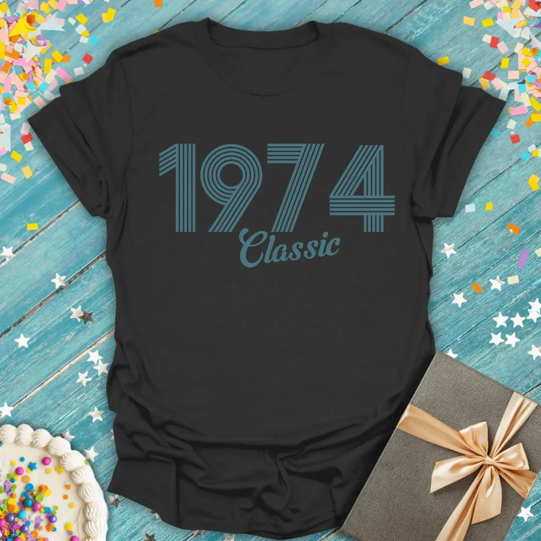 Black 1974 Classic birthday tee with confetti, cake, and gift on blue wood background