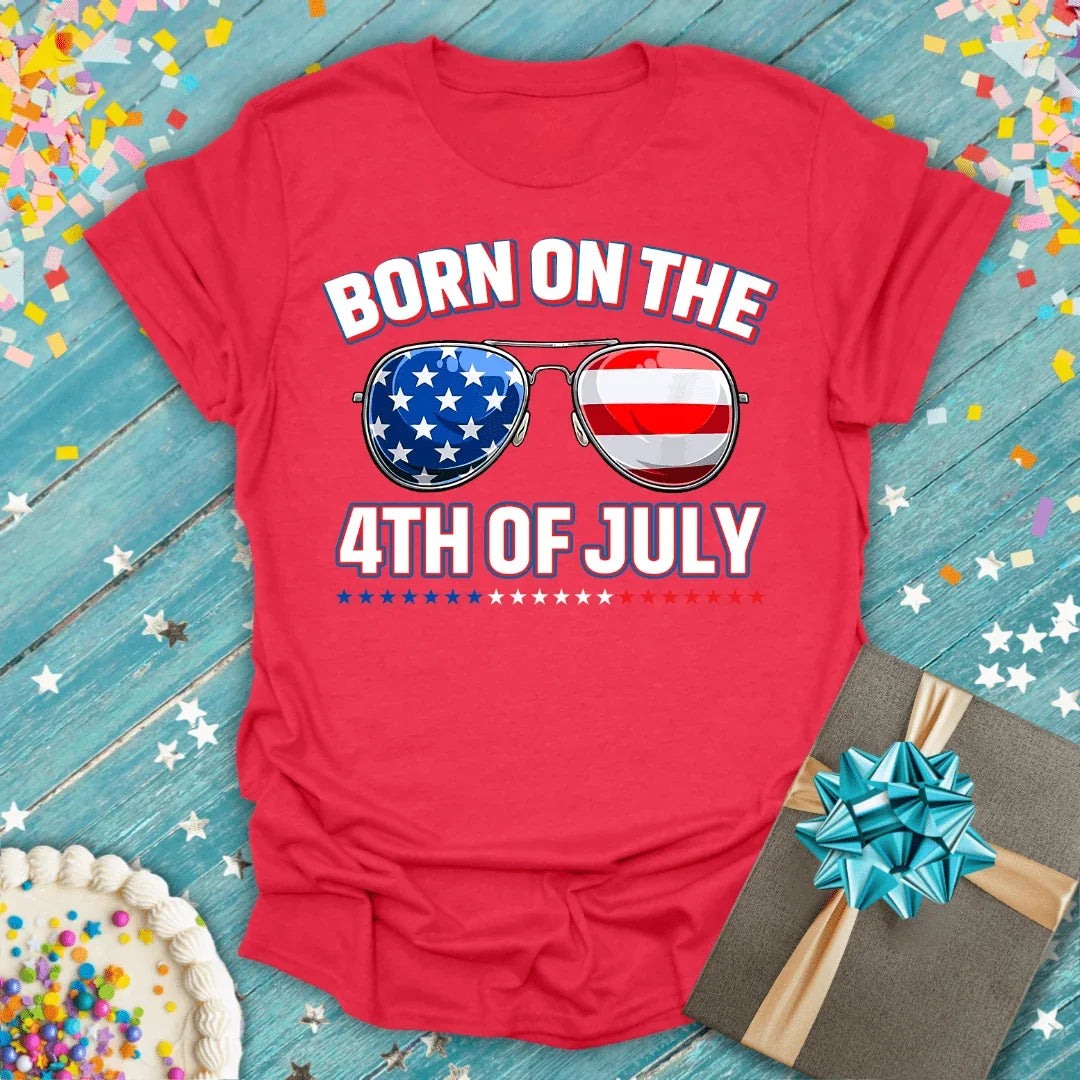 Red 4th of July birthday tee with patriotic sunglasses graphic, cake, and gift on blue wood