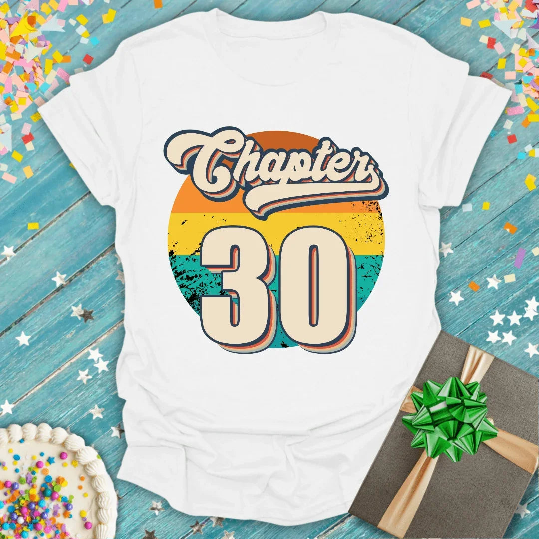 White 'Chapter 30' birthday tee with retro sunset graphic, gift box, and confetti on blue wood