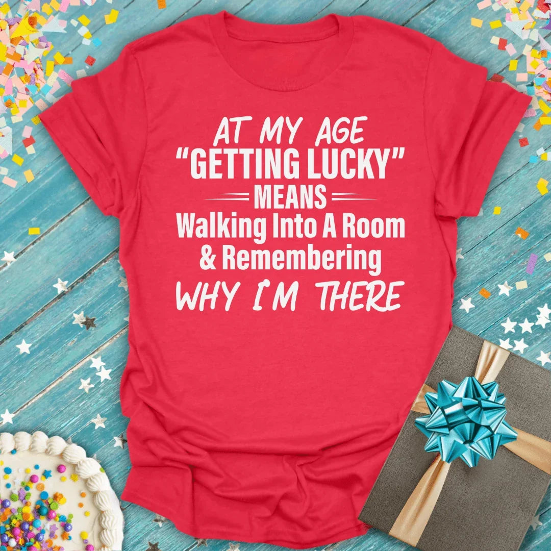 Red funny birthday tee: 'At my age getting lucky means walking into a room & remembering why I'm there' with confetti, cake, gift