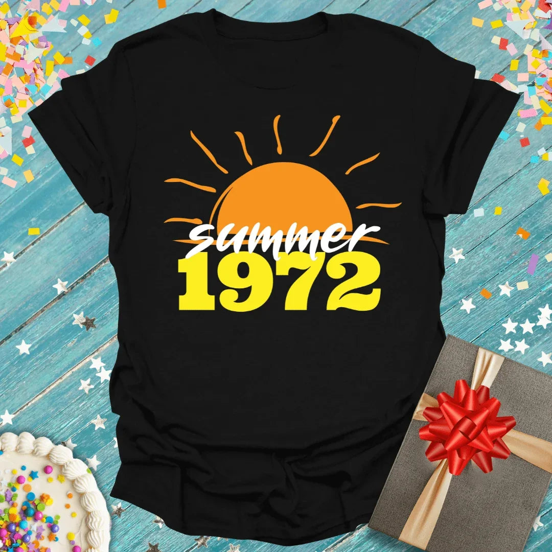 Black Summer 1972 birthday tee with sun graphic, gift box, and cake on blue wood background