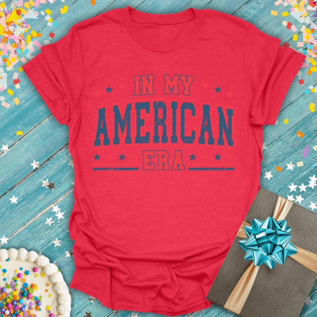 Red 'In My American Era' graphic tee with stars, gift box, and birthday cake on blue wood