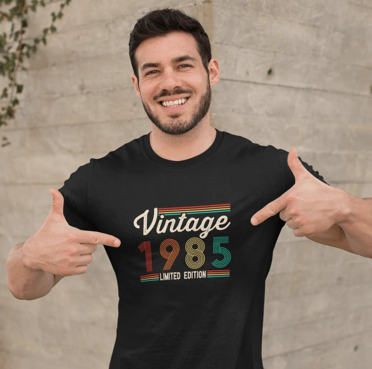 Man smiling in black vintage 1985 limited edition tee, pointing to retro design, perfect for birthdays or trips.