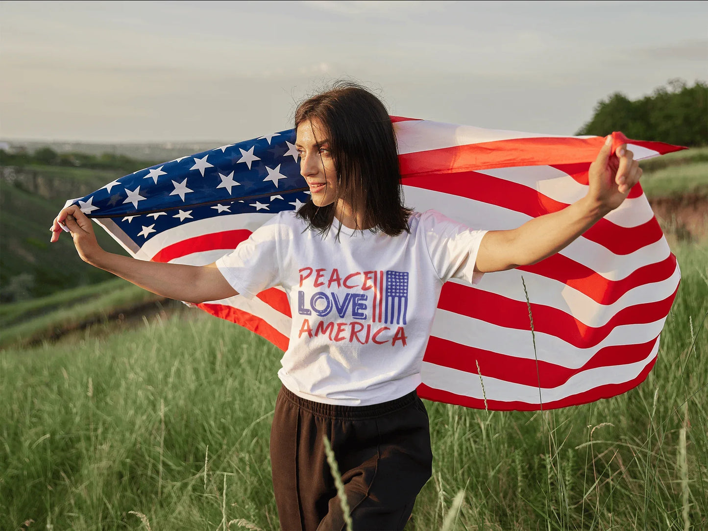 Woman in 'Peace Love America' tee holding US flag in grassy field, patriotic soft t-shirt for birthdays & trips