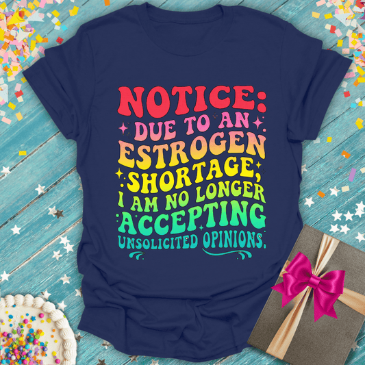 Navy blue t-shirt with colorful text 'Notice: Due to an Estrogen Shortage, I am no longer accepting unsolicited opinions' on a blue wood floor with confetti.