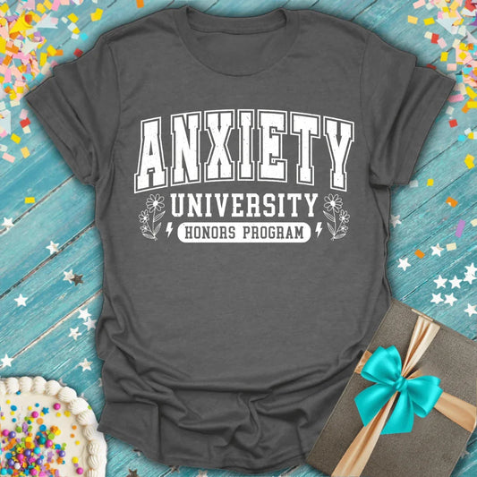Gray t-shirt with 'Anxiety University Honors Program' text on a wooden surface with confetti and a gift.