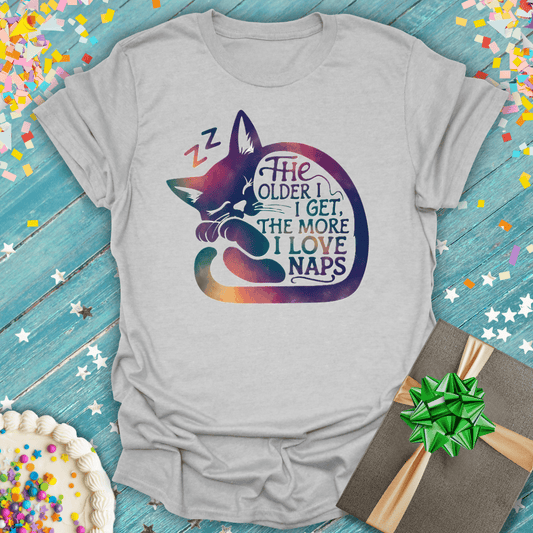 Gray t-shirt with sleeping cat graphic and quote 'The older I get, the more I love naps' on blue wood background with confetti, cake, and gift.