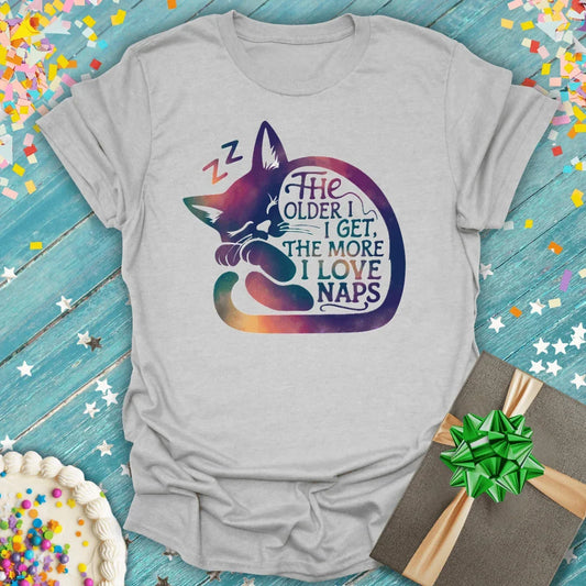 Gray t-shirt with sleeping cat graphic and quote 'The older I get, the more I love naps' on blue wood background with confetti, cake, and gift.