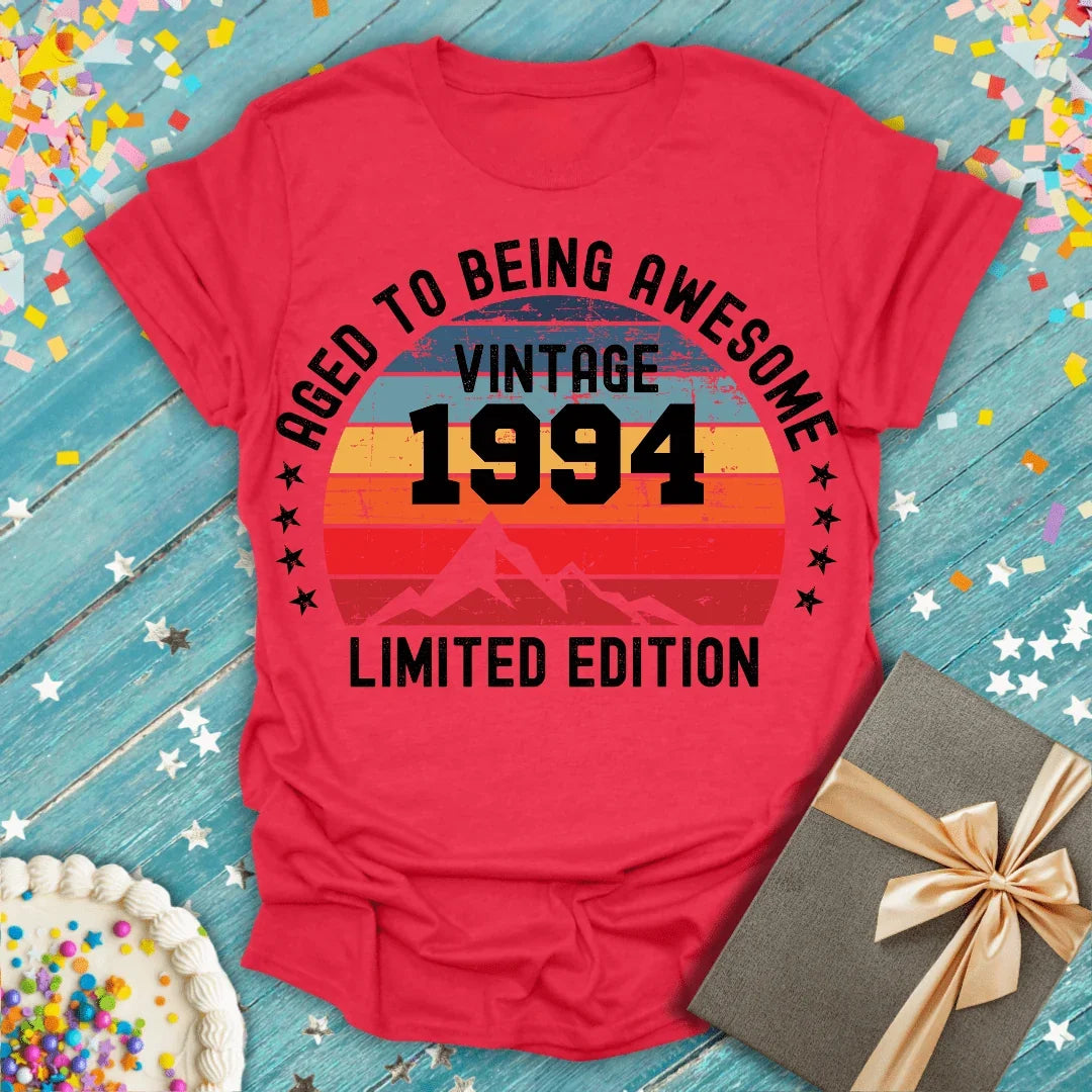 Red vintage 1994 birthday tee with 'Aged to Being Awesome' graphic, confetti, cake, and gift on blue wood