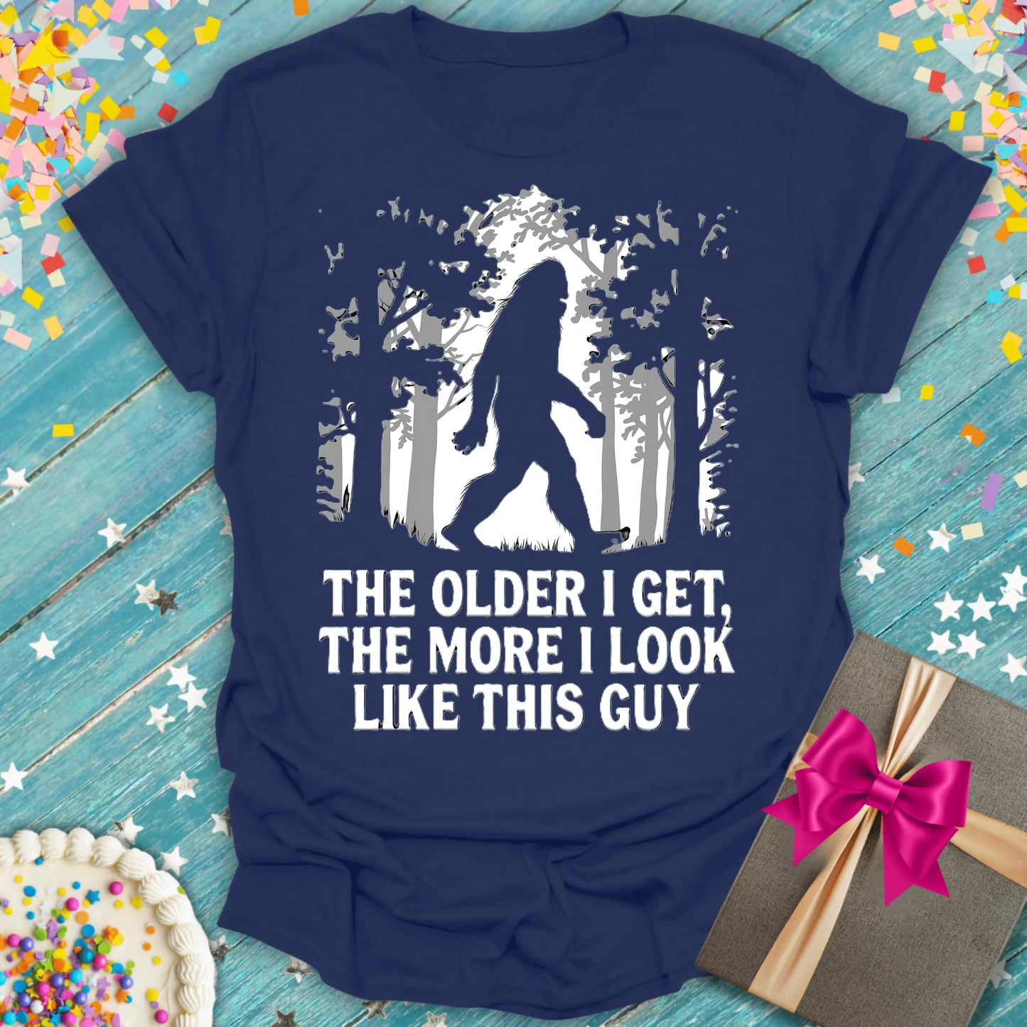 Navy t-shirt with Bigfoot graphic and text 'The older I get, the more I look like this guy' on blue wood with confetti and gift