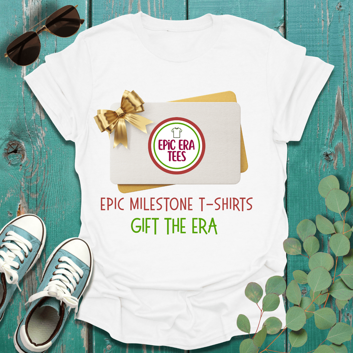 White milestone t-shirt with 'Epic Milestone T-Shirts Gift the Era' text, gift card, sunglasses, sneakers, and eucalyptus on teal wood.