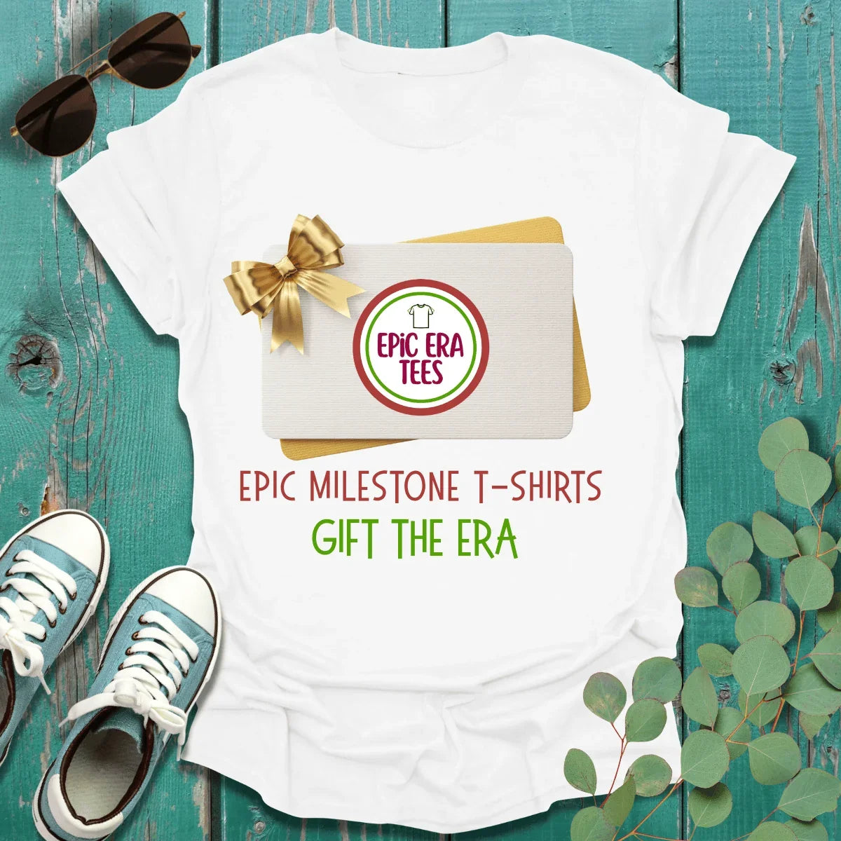 White milestone t-shirt with 'Epic Milestone T-Shirts Gift the Era' text, gift card, sunglasses, sneakers, and eucalyptus on teal wood.