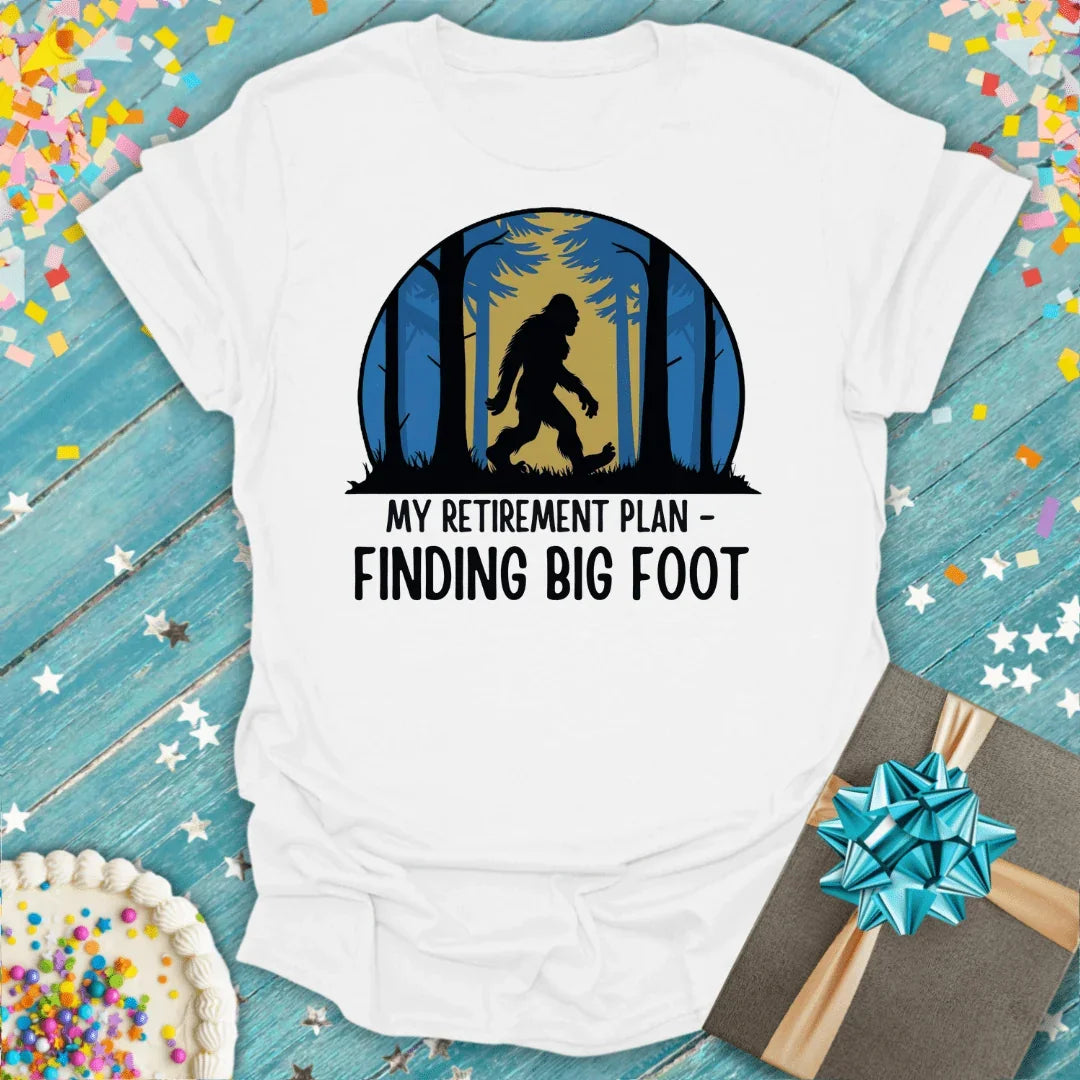White tee with Bigfoot forest design, 'My Retirement Plan - Finding Big Foot' text, gift and cake nearby