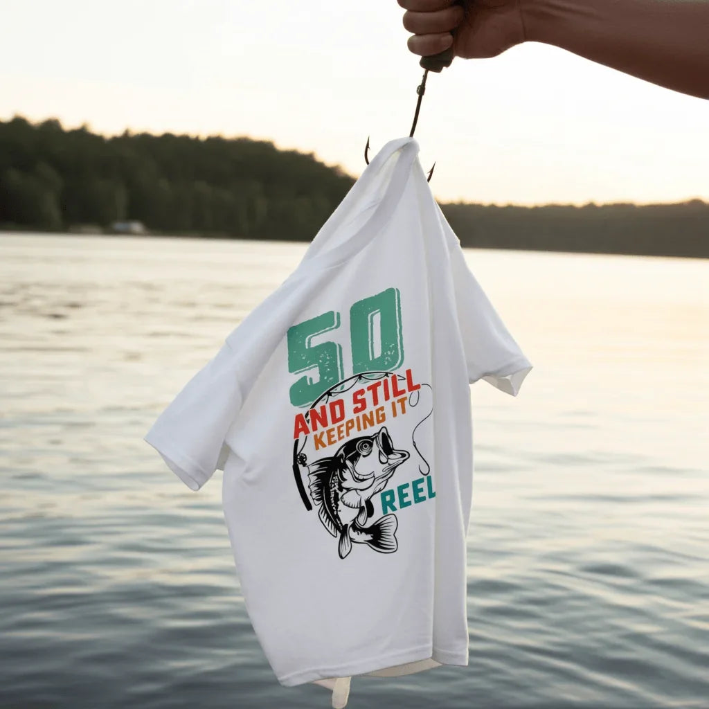 White 50th birthday fishing tee with '50 and still keeping it reel' graphic, held on hook by lake