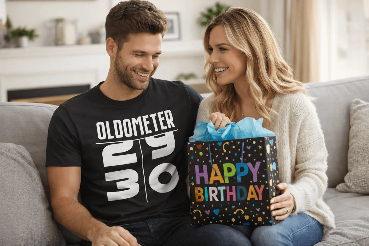 Man in 'Oldometer 29 to 30' birthday tee smiles with woman holding colorful Happy Birthday gift bag on couch