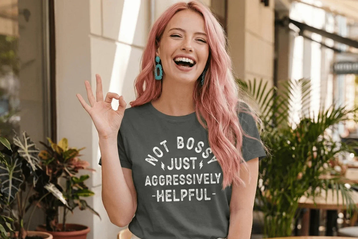 Woman in gray tee with 'Not Bossy Just Aggressively Helpful' slogan, pink hair, making OK sign outdoors