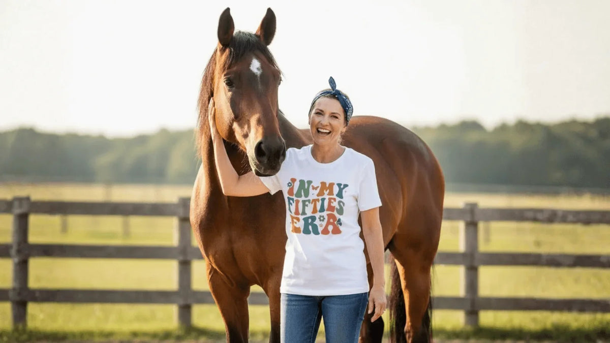 Woman in 'In My Fifties Era' tee smiling beside horse at sunset, soft birthday gift tee
