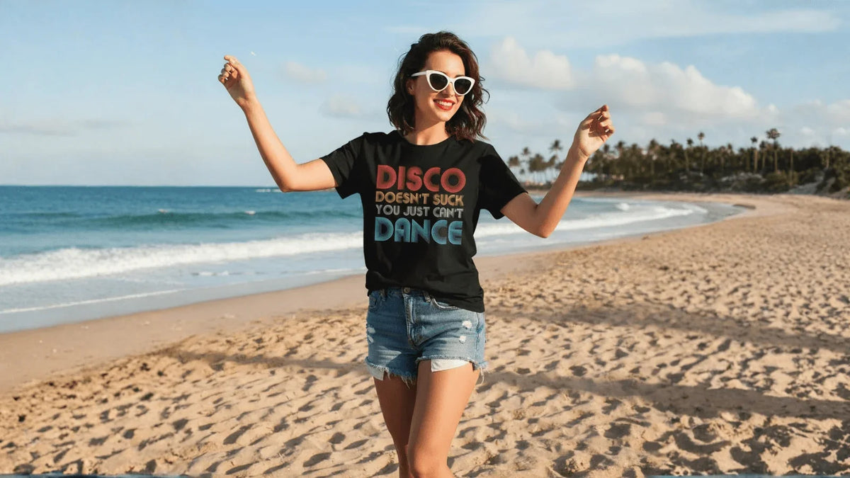 Woman dancing on beach in black 'Disco Doesn't Suck You Just Can't Dance' tee, denim shorts, white sunglasses