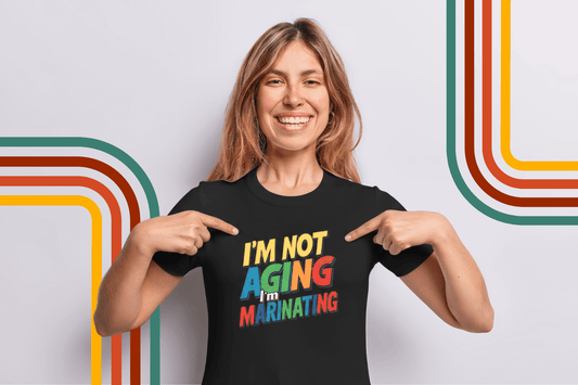 Woman smiling in black tee with colorful 'I'm not aging, I'm marinating' text, pointing at design