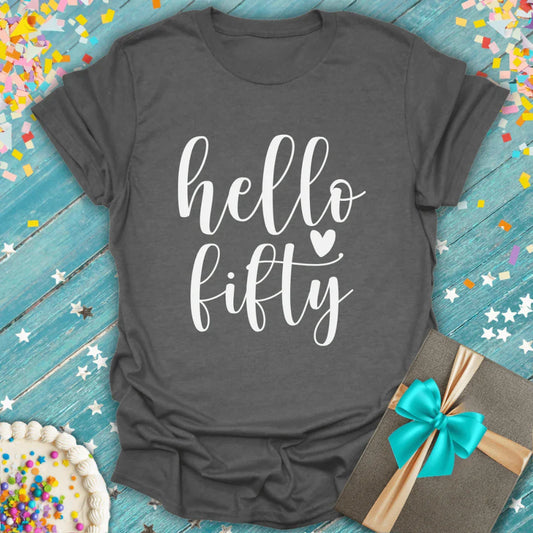 Gray birthday T-shirt with 'hello fifty' text, confetti, cake, and gift on blue wood background