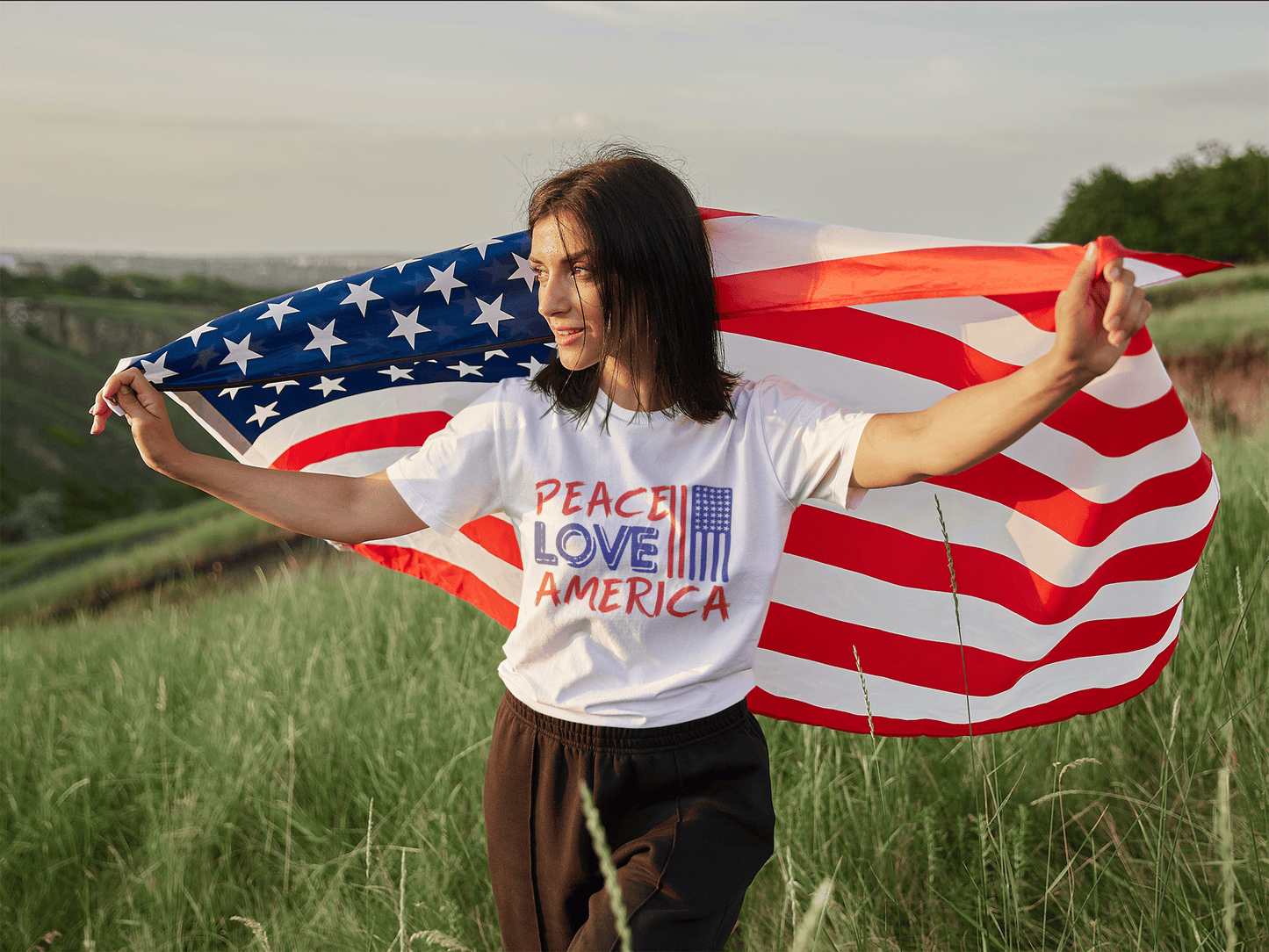 Woman in 'Peace Love America' tee holding US flag in grassy field, patriotic soft t-shirt for birthdays & trips
