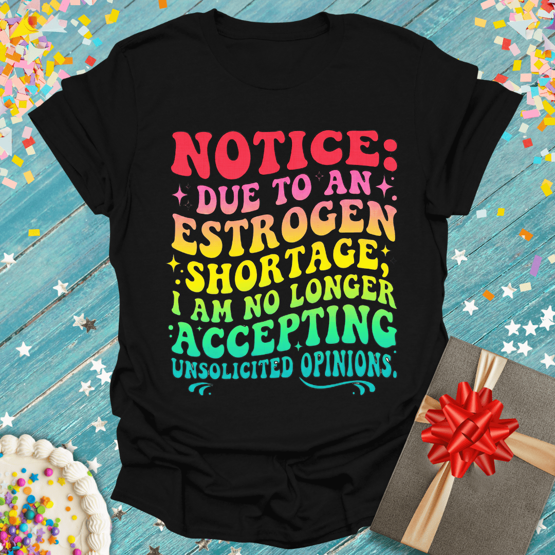 Black funny birthday tee with colorful 'Estrogen Shortage' quote, gift box & cake on blue wood