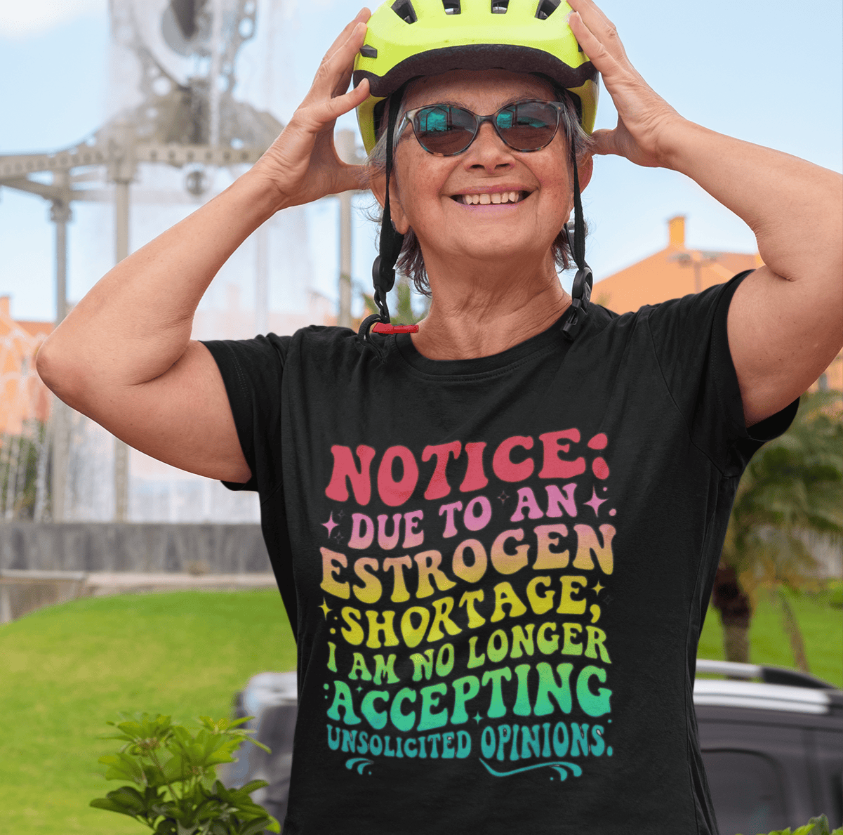 Woman in black tee with bold text 'Notice: Due to an estrogen shortage...' adjusting yellow bike helmet outdoors