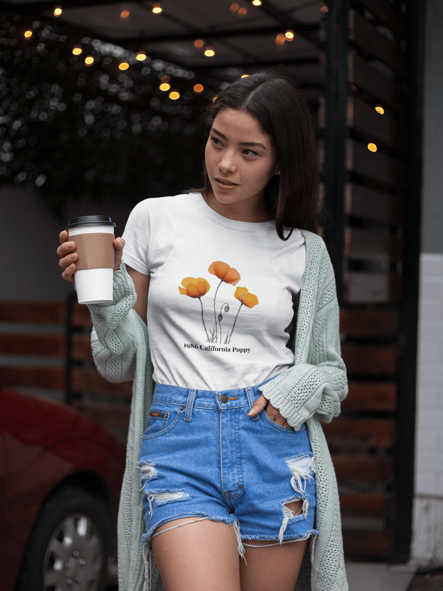 Woman in white 1986 California Poppy tee, denim shorts, holding coffee, outdoor cafe vibe