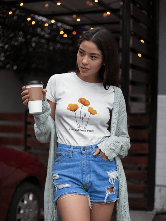 Woman in white 1986 California Poppy tee, denim shorts, holding coffee, outdoor cafe vibe