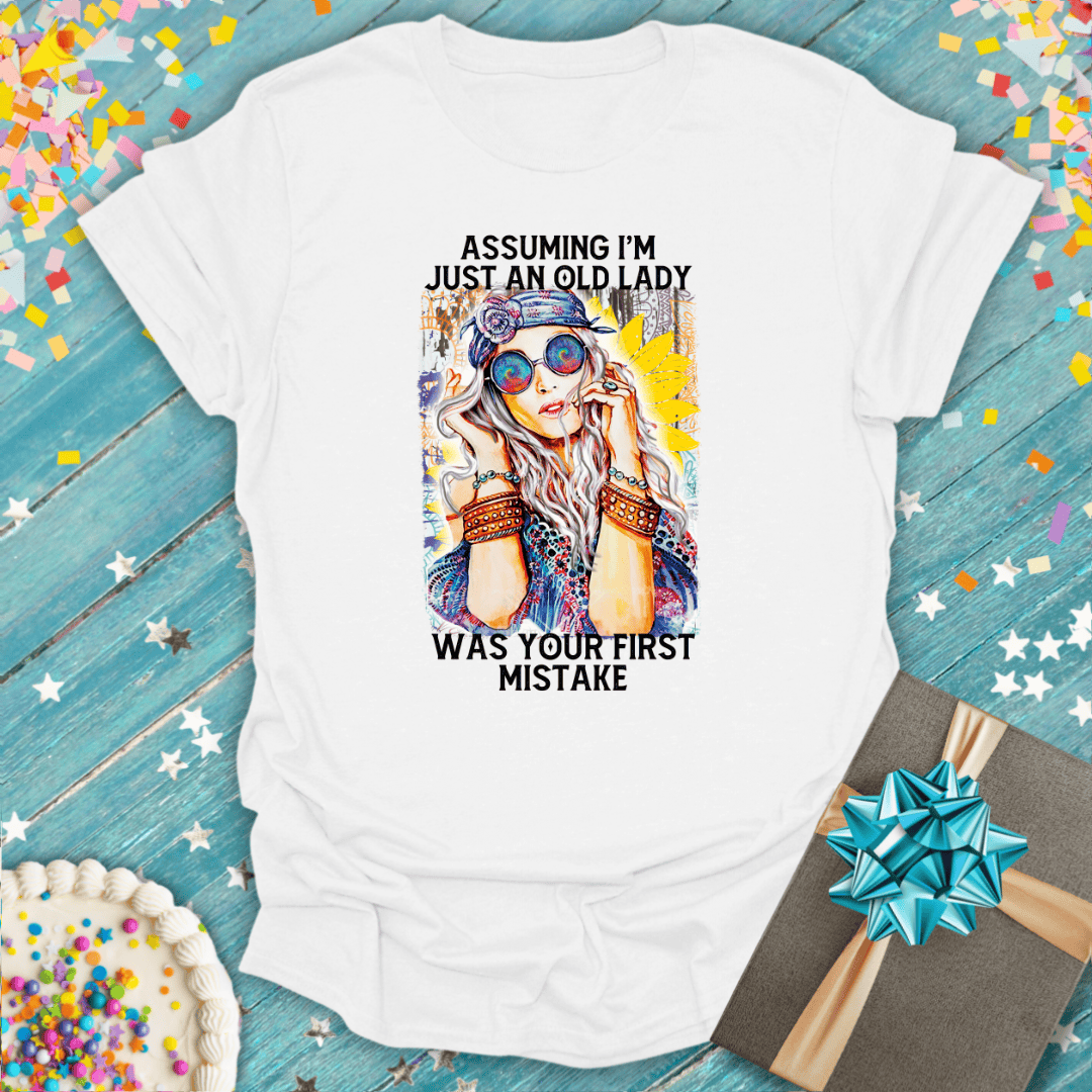 White graphic tee with 'Assuming I'm just an old lady was your first mistake' text, colorful confetti, cake, and gift