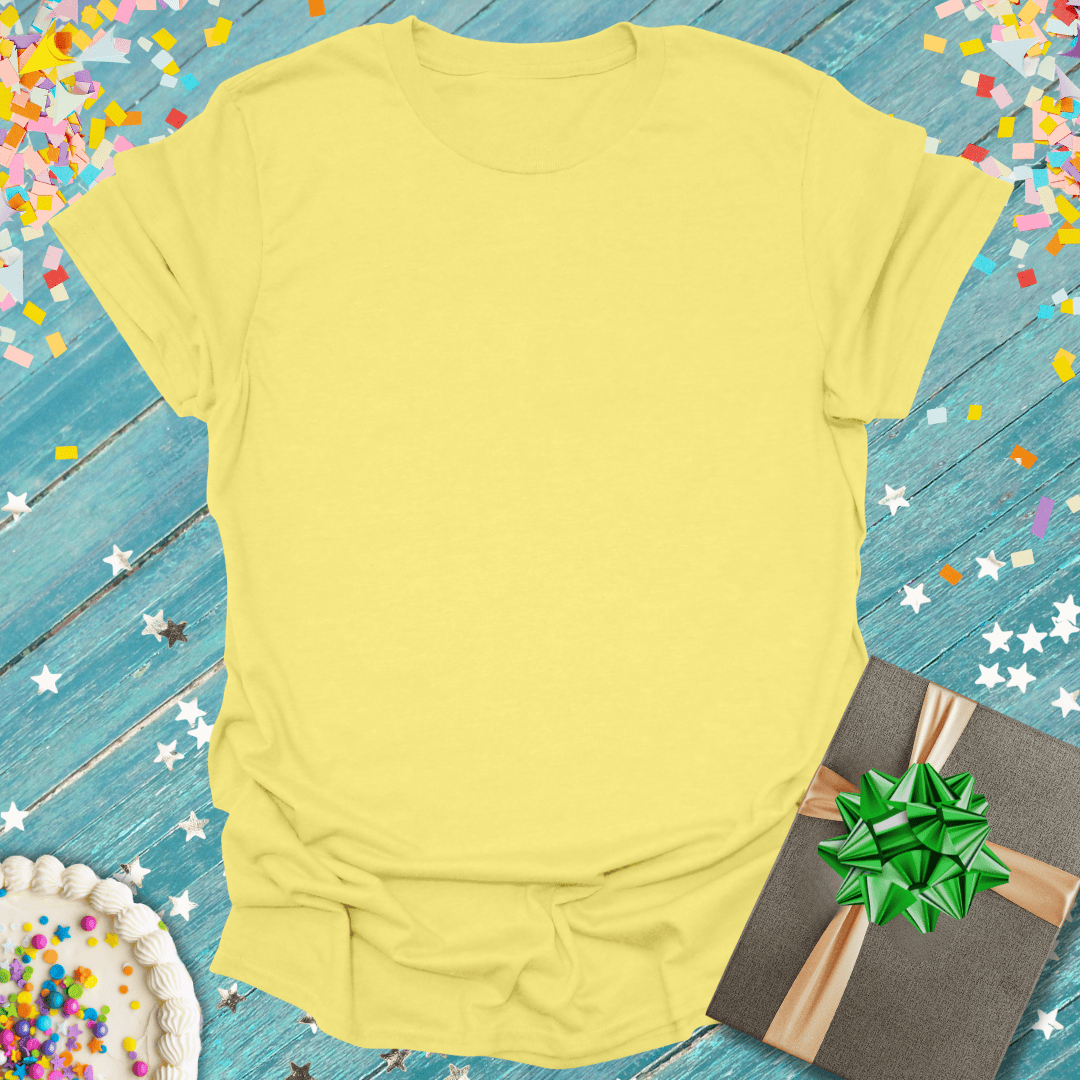 Soft yellow tee with birthday cake, gift box, and confetti on blue wood — perfect for celebrations and trips.
