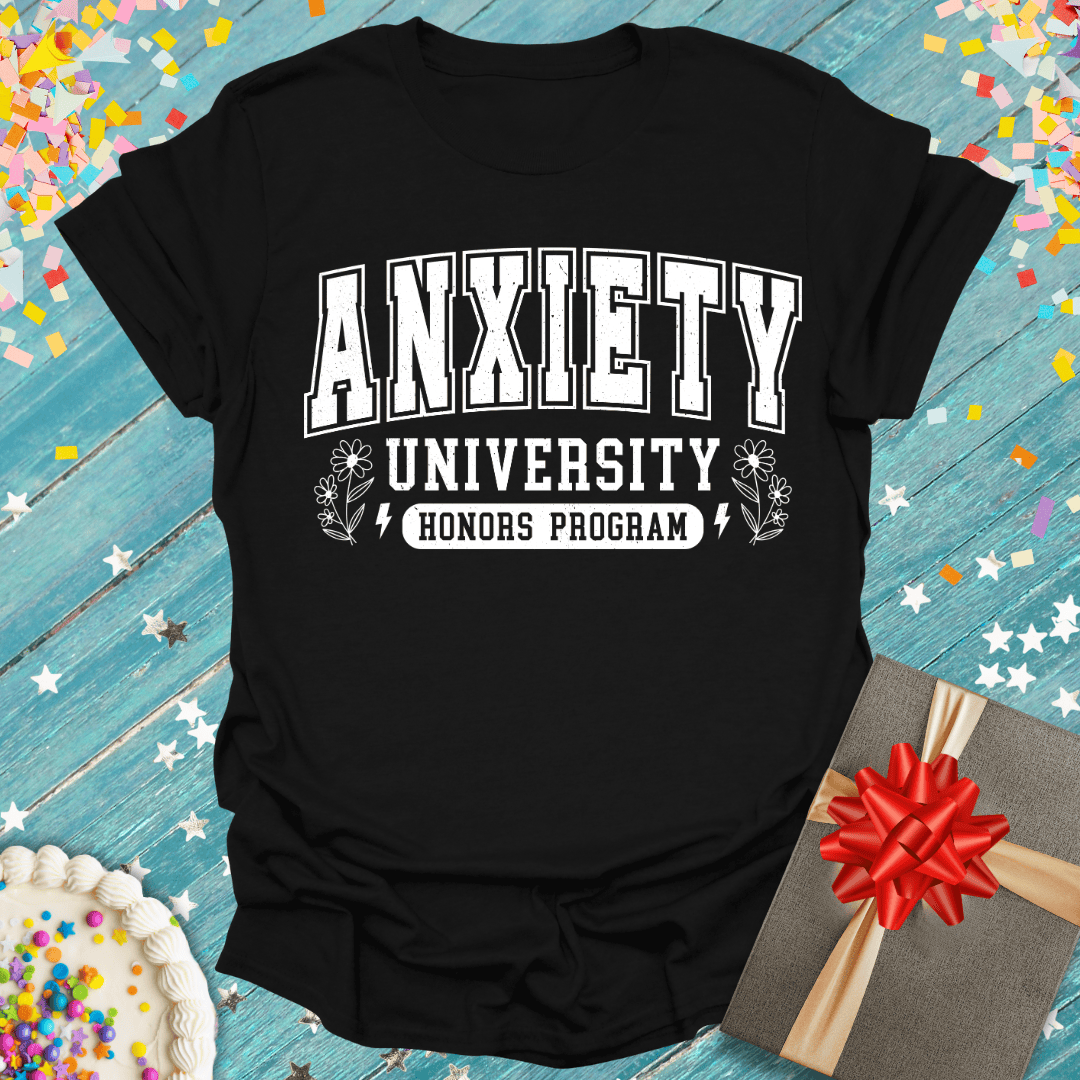 Black t-shirt with 'Anxiety University Honors Program' text on a blue wooden surface with confetti and a gift.