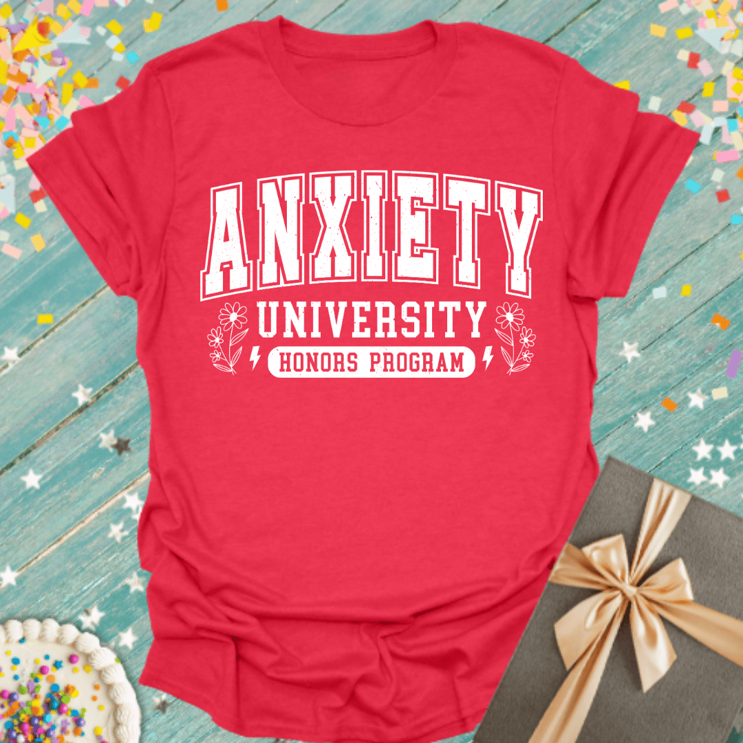 Red t-shirt with 'Anxiety University Honors Program' text on a blue wooden surface with confetti and a gift.