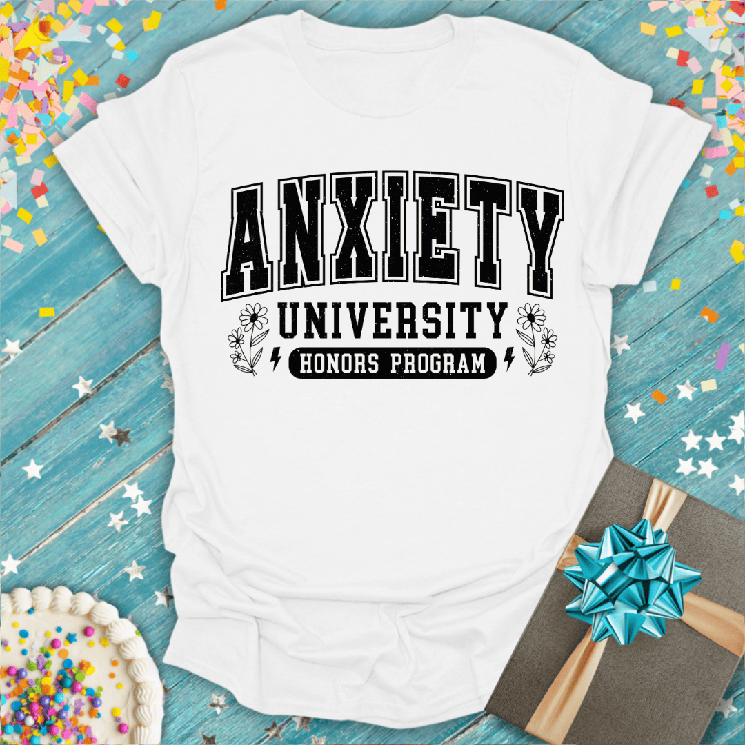 White t-shirt with 'Anxiety University Honors Program' text on a blue wooden surface with confetti and a gift.