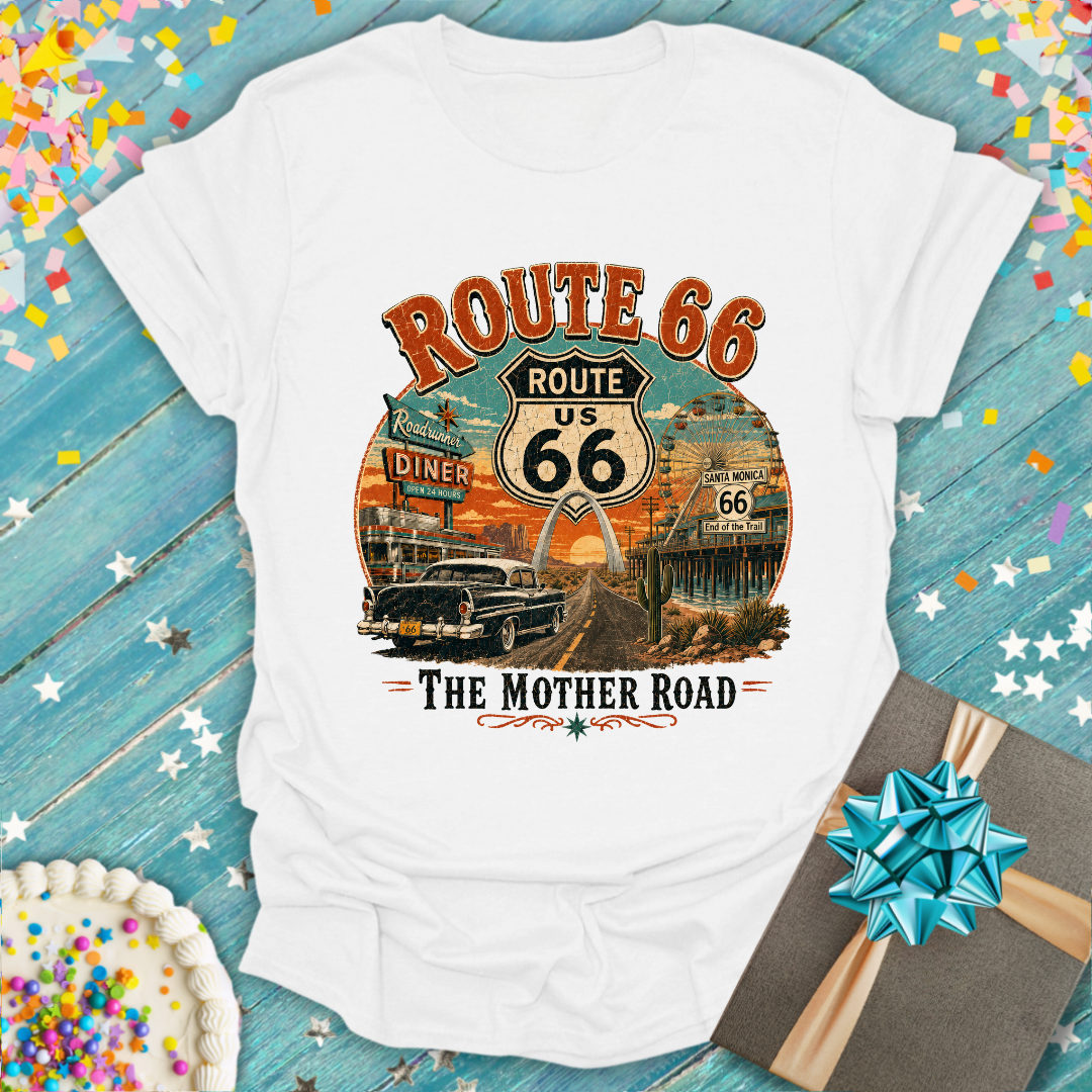 A white short-sleeve graphic T-shirt laid flat on a rustic blue wooden surface with confetti and a gift box nearby. The shirt features a large vintage-style Route 66 graphic with orange lettering, a central shield sign reading US 66, retro diner signs, a classic car, and the words 'The Mother Road'.