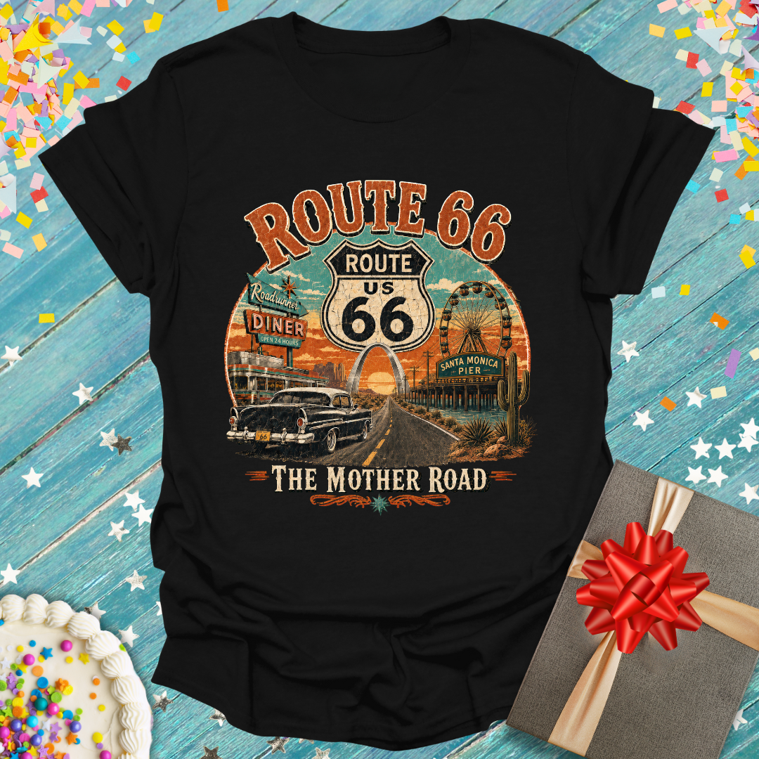 A black short-sleeve graphic T-shirt laid flat on a rustic blue wooden surface with confetti and gift props. The shirt features a large vintage-style Route 66 print with orange, white, and tan tones, including text reading 'ROUTE 66', 'US 66', and 'THE MOTHER ROAD' along with illustrated cars, signs, and a sunset scene.