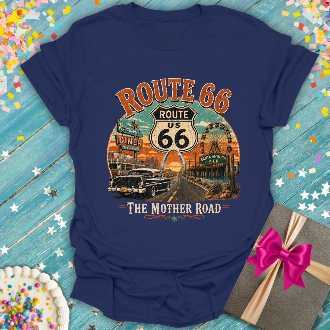 A navy blue graphic t-shirt featuring a retro 'Route 66 The Mother Road' design, laid flat on a rustic blue wooden background with confetti, a cake, and a gift box.
