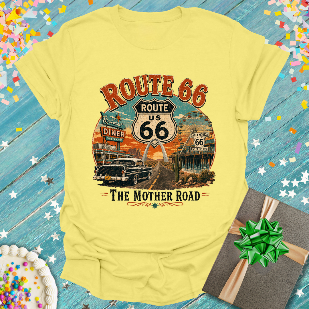 A pale yellow graphic T-shirt laid flat on a rustic blue wooden surface with confetti and a gift box around it. The shirt features a large vintage-style Route 66 graphic with the words 'ROUTE 66 US' and 'THE MOTHER ROAD' plus an illustrated car, diner, and sunset scene.