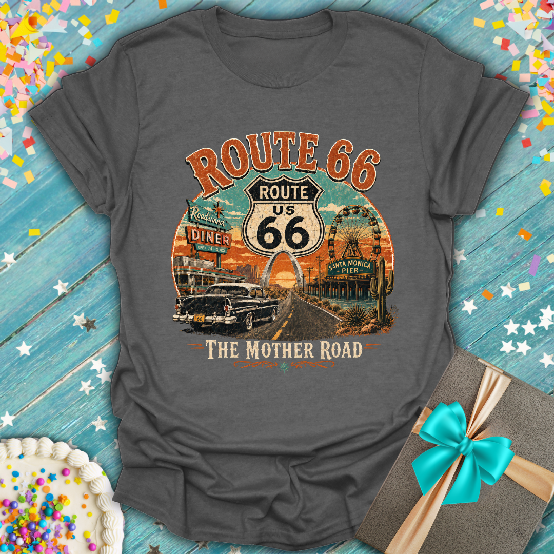 A dark gray graphic t-shirt featuring a vintage-style 'Route 66 The Mother Road' print with a retro car, diner, and desert scenery. The shirt is displayed flat on a rustic blue wooden background decorated with confetti, a birthday cake, and a gift box.