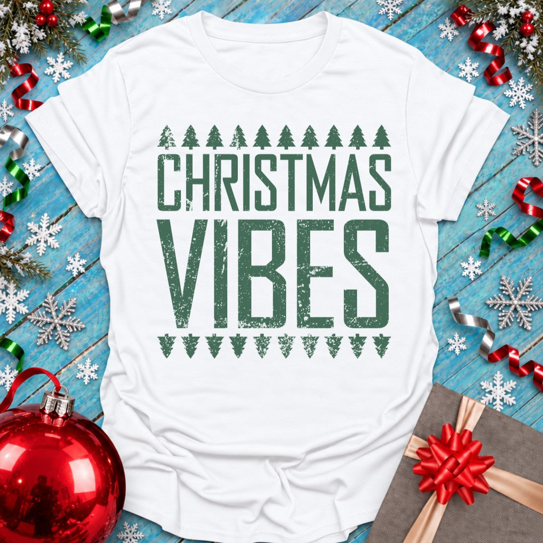 A white short-sleeve T-shirt laid flat on a blue wooden surface, printed with the words “CHRISTMAS VIBES” in distressed dark green lettering, flanked by small Christmas tree silhouettes. The shirt is styled with holiday decorations including a red ornament, red gift bow, silver snowflakes, green and red ribbons, and a wrapped gift box.