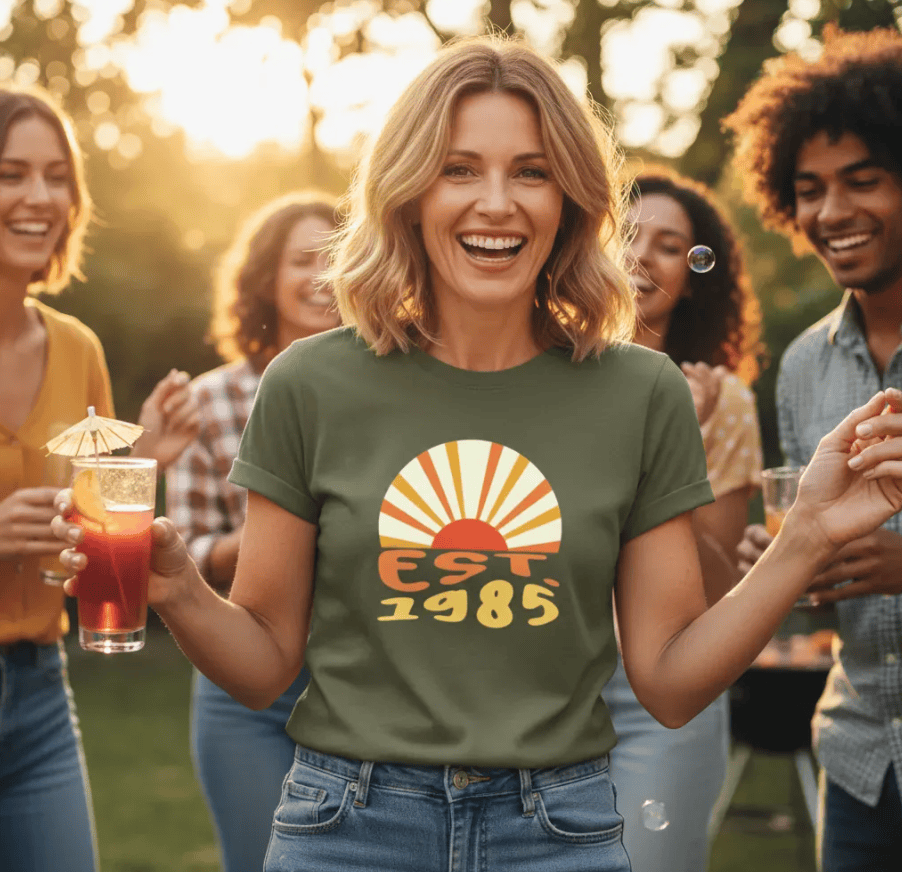 Woman in olive green 'EST. 1985' sunset tee at outdoor party, holding drink, celebrating with friends