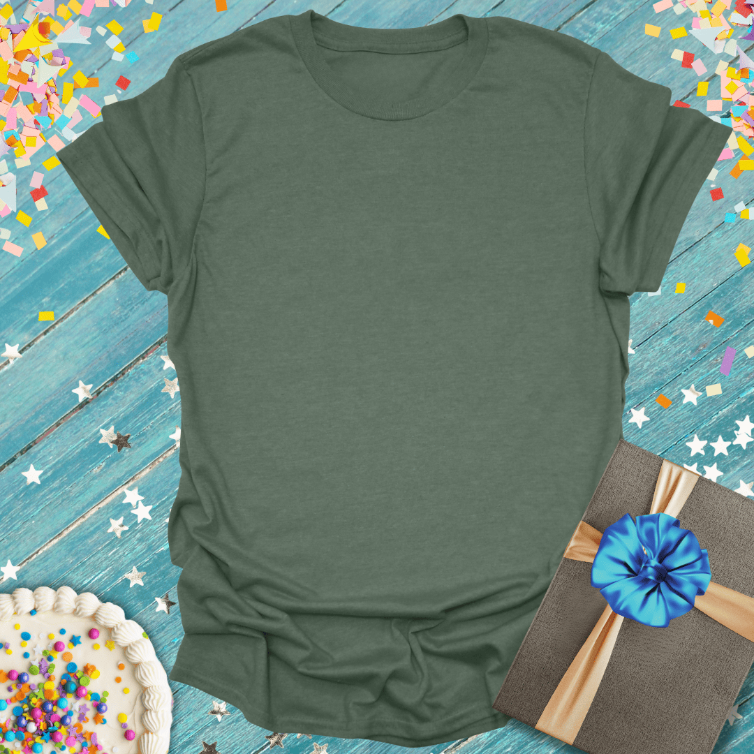 Soft olive green tee with birthday cake, gift box, and confetti on blue wood — perfect for celebrations and trips.