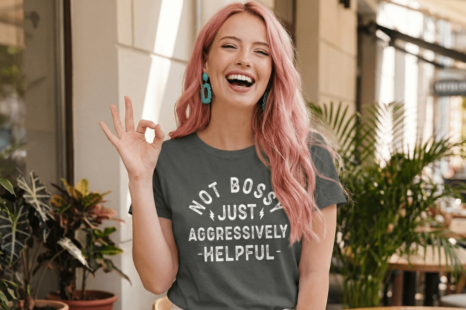 Woman in gray tee with 'Not Bossy Just Aggressively Helpful' slogan, pink hair, making OK sign outdoors