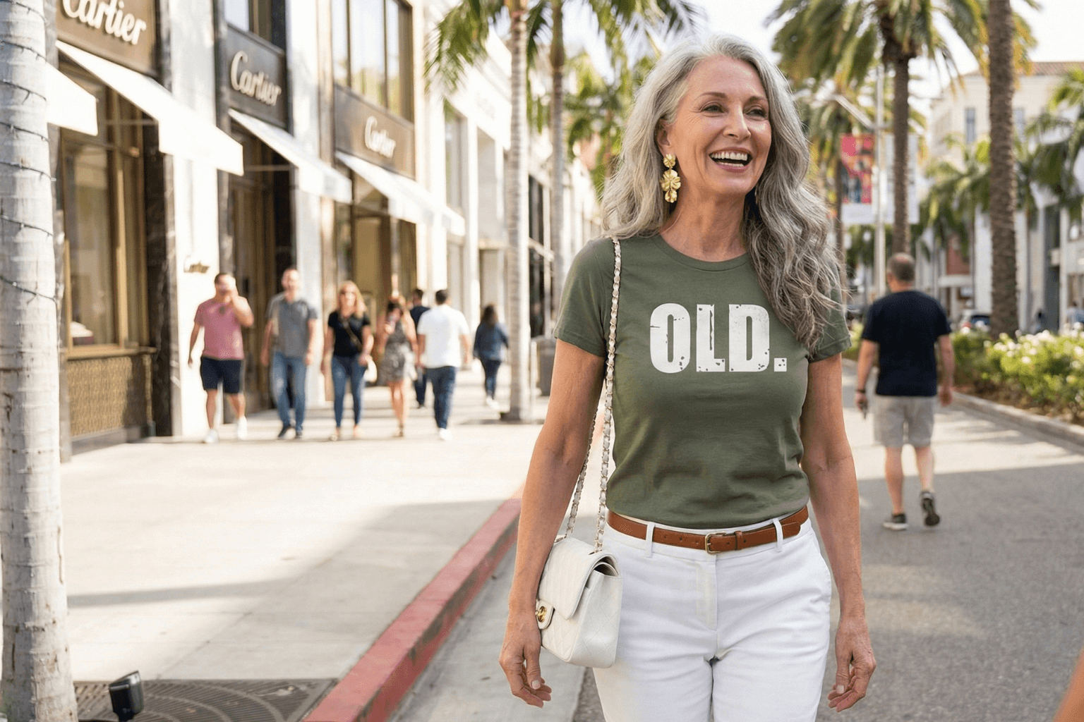 Woman wearing olive green 'OLD.' graphic tee, white pants, walking on palm-lined street